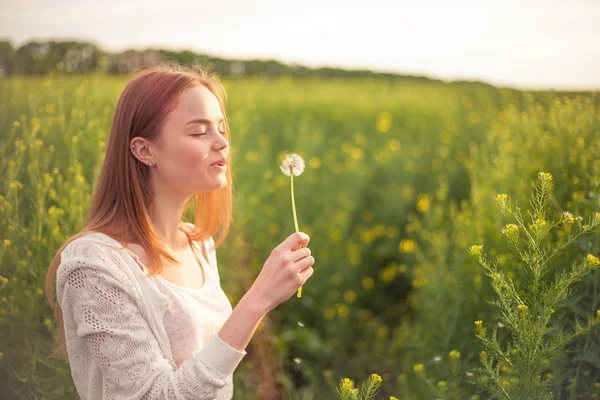 Joven mujer de la moda de primavera soplando diente de león en el jardín de primavera. Primavera. Chica de moda al atardecer en el fondo del paisaje de primavera —  Fotos de Stock