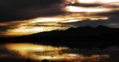 Dağların üzerinde turuncu gün batımı ve gölde yansımalar. 