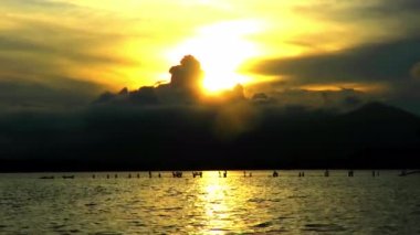 Bulutlarla kaplı dağların üzerinde turuncu gün batımı ve göldeki kuşların siluetleriyle yansımalar.