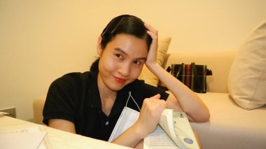 Asyalı bir kadın gülümsüyor ve kameraya bakıyor, evde oturmuş kitap okuyor.