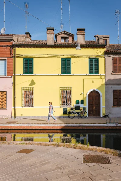 Comacchio, İtalya 'nın merkezinde, kanalın kıyısında yürüyen bir kadın arkasında sarı bir ev var..