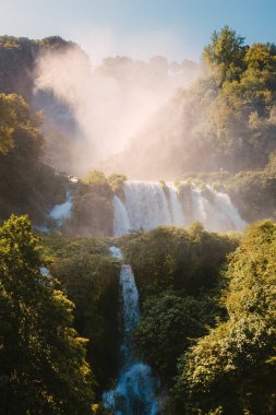 Marmore, İtalya - 19 Ağustos 2025: Güneş ışığı, orman tepe örtüsü üzerinde yükselen görünür sis bulutlarıyla şelaleyi aydınlatıyor.