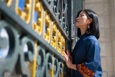 Genç Asyalı kadın model geleneksel elbiseyle poz veriyor ve arka planda imparatorluk çiti var.