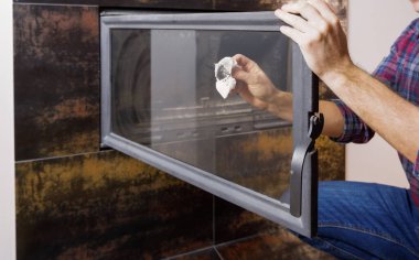 Gömlekli ve kot pantolonlu bir adam modern bir şöminenin ısıya dayanıklı camını temizliyor. Ellerin yakın görüntüsü.