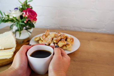 Bir fincan kahve kadınların elinde, yakın plan. Karantina sırasında ev yapımı pişirilmiş ürünler. Ev yapımı tatlı hazırlığı. Tahta bir masanın üstünde ev yapımı beyaz peynir. Beyaz tuğla bir duvar ve bir buket gülün arka planında kavram.