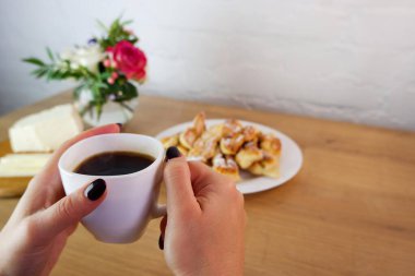 Bir fincan kahve kadınların elinde, yakın plan. Karantina sırasında ev yapımı pişirilmiş ürünler. Ev yapımı tatlı hazırlığı. Tahta bir masanın üstünde ev yapımı beyaz peynir. Beyaz tuğla bir duvar ve bir buket gülün arka planında kavram.