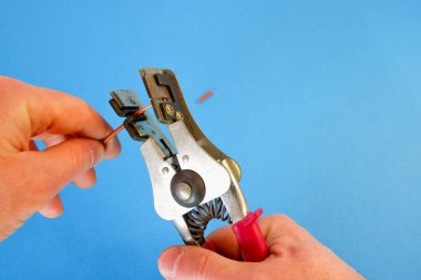 Elektrikçi özel bir striptizci aleti kullanarak kablodaki yalıtımı kaldırıyor. Mavi bir arka planda elleri yakın çekim.