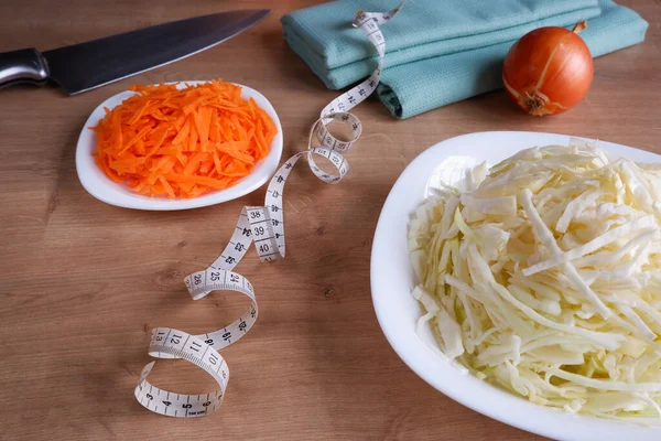 Healthy eating. Raw, chopped vegetables for salad on white plates. Cabbage, carrots, onions, measuring tape on a wooden table