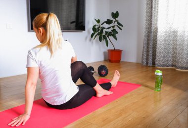 Ev antrenmanı. Siyah taytlı, tanınmayan sarışın kadın evdeki pembe paspasın üzerinde halterlerle güç antrenmanı yapıyor. Kablosuz kulaklık ve su şişesi..