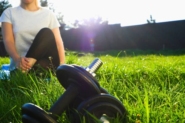 Outdoor sports. Dumbbells on green grass in the backlight of the setting sun. The woman in the background stretches after a hard workout.