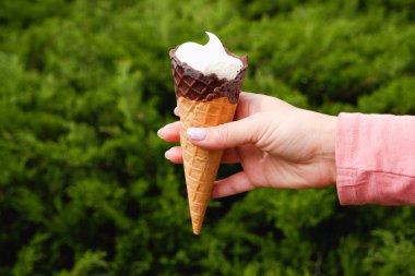 Manikürlü bir kadının elinde yeşil bir doğa parkının arka planında çikolatalı waffle külahı vardır..