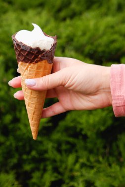 Manikürlü bir kadının elinde yeşil bir doğa parkının arka planında çikolatalı waffle külahı vardır..