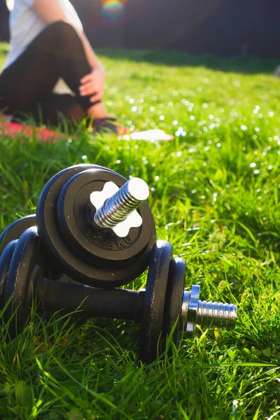 Outdoor sports. Dumbbells on green grass in the backlight of the setting sun. The woman in the background stretches after a hard workout.