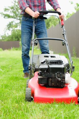 Ekose gömlekli ve kot pantolonlu bir adam çimleri çim biçme makinesiyle biçiyor..