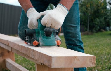 Marangozlar eldivenleri, elektrikli uçakları, keresteleri, uçan talaşları, talaşları. Eldivenli ellerde elektrikli bir uçakla yakın plan..