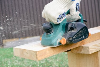 Marangozlar eldivenleri, elektrikli uçakları, keresteleri, uçan talaşları, talaşları. Eldivenli ellerde elektrikli bir uçakla yakın plan..