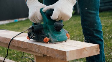 Marangozlar eldivenleri, elektrikli uçakları, keresteleri, uçan talaşları, talaşları. Eldivenli ellerde elektrikli bir uçakla yakın plan..