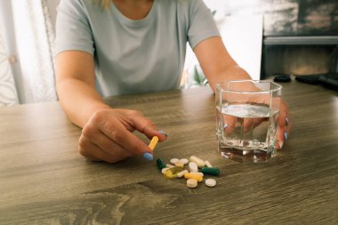 Elinde farklı haplar ve bir bardak su olan arka planda takviye şişeleri olan bir kadının yakın çekimi. Sağlık, ilaç, vitamin, tedavi ve günlük destek maddeleri kavramı.