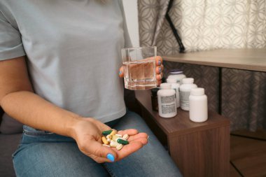 Elinde farklı haplar ve bir bardak su olan arka planda takviye şişeleri olan bir kadının yakın çekimi. Sağlık, ilaç, vitamin, tedavi ve günlük destek maddeleri kavramı.