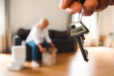 Yakın plan bir el, ev ya da dairenin anahtarlarını net bir şekilde tutuyor. Bulanık arka planda, bir kişi bir kanepede oturuyor, parlak, modern bir oturma odasındaki kutuları açıyor..
