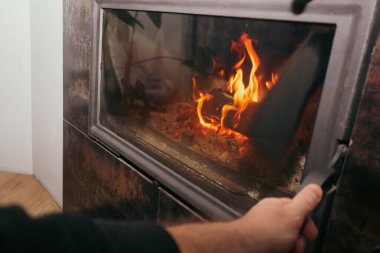 Cam şömine kapısının kulpunu tutan birinin yakın çekimi. Kışın şöminenin içinde parlak turuncu alevler ve parlayan közler görülebilir..
