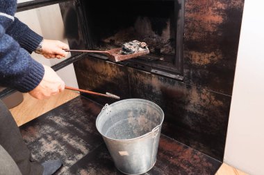 Kazaklı yaşlı bir adam soğuk kül ve kurumu açık modern bir şömineden galvanizli metal kovaya çıkarıyor. Isınma mevsiminde rutin bakım ve temizlik.