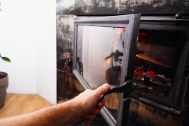 Cam şömine kapısının kulpunu tutan birinin yakın çekimi. Kışın şöminenin içinde parlak turuncu alevler ve parlayan közler görülebilir..