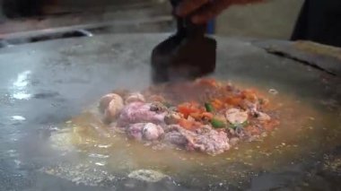 Katakat Baharatlı Et sıcak bir ızgarada cızırdıyor, buhar yükseliyor, dışarıda lezzetli sokak yemekleri sergiliyor.