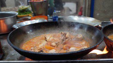 Close-up of chicken karahi in a wok over flames, steam rising, with cooking equipment around, street food