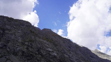 Parlak mavi gökyüzü ve kabarık bulutlara karşı sarp kayalık bir dağ yüzeyinin dramatik fotoğrafı, heybetli bir kavram..