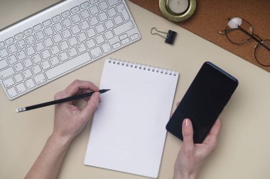 Düz yatış, dizüstü bilgisayarı, klavyesi, not defteri, telefonu, kalemi, kalemi, gözlüğü ve yeşil bitkisi olan ofis masası. Fotokopi arkaplanı olan bir solak elinde kalem, bir kalem tutuyor. Telefonunu elinle tut.