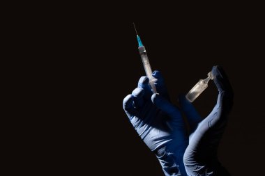 Siyah arka planda, mavi eldivenli eller bir şırınga ve bir ampul aşı tutuyor. Coronavirus, karantina, tecrit, aşı. Tıbbi konsept.