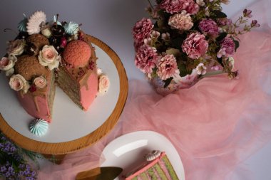 Kremalı kurabiyelerle süslenmiş pembe bir doğum günü pastası. Yanında pembe çiçeklerden oluşan bir vazo var. Şekerleme ürünleri.