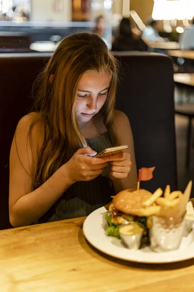 Önünde bir tabak yemekle bir restoranda telefonunu kullanan 12 yaşında sarışın bir beyaz kız..