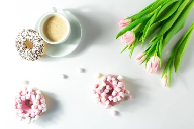 Cappuccino ve marşmelovlu donutlar beyaz arka planda, pembe laleler üst tarafta, izole edilmiş.