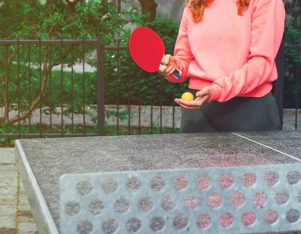 Pembe kazaklı bir kız dışarıda tenis masasında pinpon oynuyor.