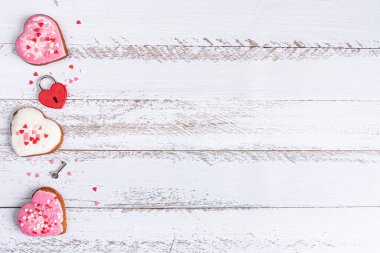 Pembe ve beyaz kalp şeklinde zencefilli ekmek, kalp şeklinde kırmızı asma kilit ve beyaz ahşap arka planda küçük kırmızı, pembe ve beyaz kalplerle çevrili bir anahtar.