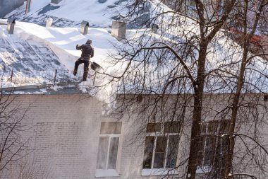 Evin çatısından kar döken bir adam yakınlardaki ağaçlardan...