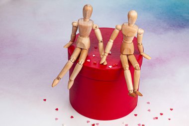 İki ahşap manken el ele tutuşuyor ve üzerinde beyaz, pembe ve kırmızı kalpler olan küçük kalpleri olan yuvarlak kırmızı bir hediye kutusunun kapağında oturuyorlar.