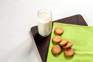 a tall glass of milk, cookies and a green towel on dark cutting board on white background