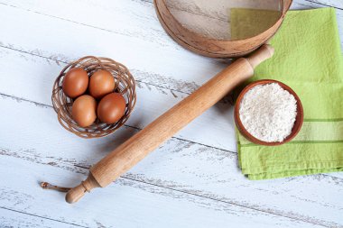 Tahta merdane, yumurta dolu küçük ahşap sepet, un dolu ahşap kase, süzgeç ve beyaz arka planda yeşil havlu.