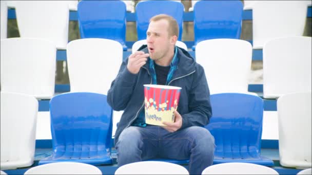 Un tipo se sentó en la tribuna con palomitas de maíz cubo — Vídeos de Stock