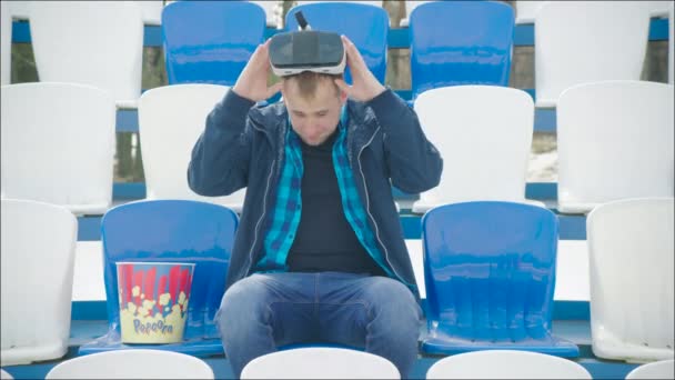 Un tipo con gafas VR y palomitas sentado en la tribuna — Vídeos de Stock