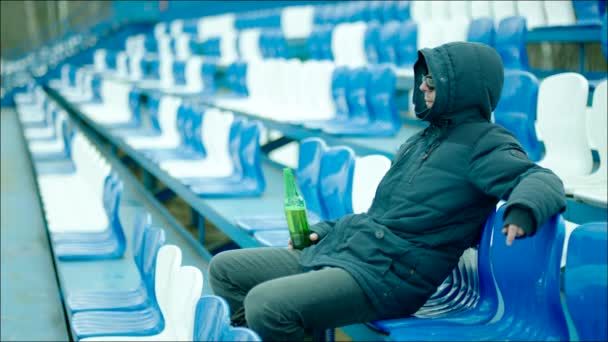 Guy in the hood sitting on the tribune and drinking from the bottle — Stock Video