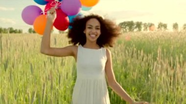 Young girl running through a wheat field with colour balloons during sunset
