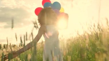 Young girl walking through a wheat field with colour balloons during sunset