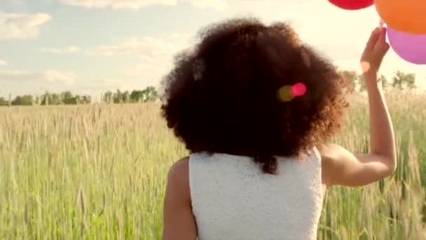 Niña corriendo a través de un campo de trigo con globos de color durante la puesta del sol — Vídeo de stock