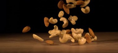 Badem ve kaju fıstıklarının süper yavaş çekimi ahşap masaya düşer. Sağlıklı atıştırmalıklar, tatmin edici çıtırtılar. Ticari siyah arkaplan