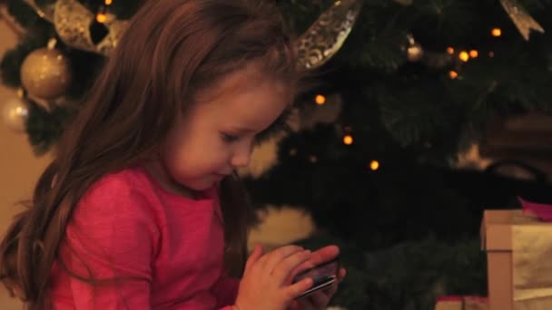 Маленькая девочка со смартфоном у елки — стоковое видео