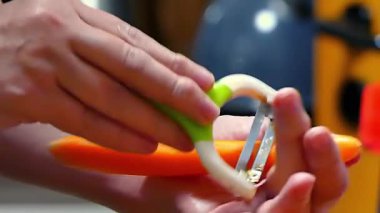 Temiz beyaz arka planda metal kabuklu taze, canlı turuncu bir havucu soyan ellerin yakın çekimi. Sağlıklı beslenme, yemek pişirme ve yemek hazırlama kavramı.